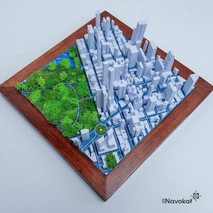 This photo features a detailed 3D miniature map of Manhattan's Central Park, showcasing lush green trees and pathways surrounded by a white cityscape of tall buildings, mounted on a sleek wooden base. The map highlights New York City's iconic urban and green spaces, making it a stylish modern wall art piece. The wooden frame adds a touch of elegance, perfect as a Manhattan-themed gift or unique home decor for city lovers.