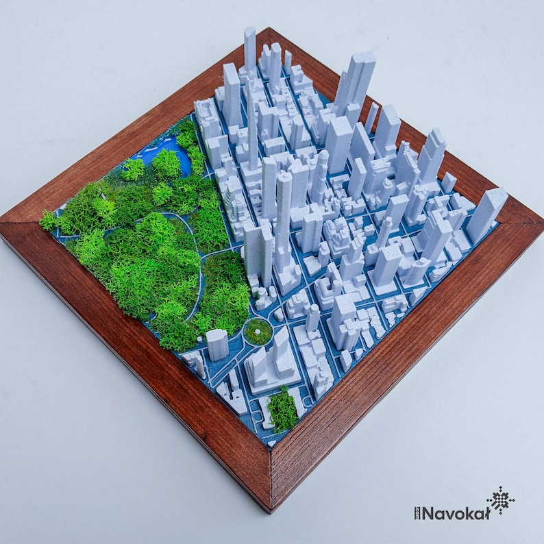 This photo features a detailed 3D miniature map of Manhattan's Central Park, showcasing lush green trees and pathways surrounded by a white cityscape of tall buildings, mounted on a sleek wooden base. The map highlights New York City's iconic urban and green spaces, making it a stylish modern wall art piece. The wooden frame adds a touch of elegance, perfect as a Manhattan-themed gift or unique home decor for city lovers.