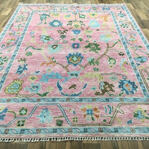 Könnte beinhalten: Ein rechteckiger, rosa Teppich mit floralem Muster. Er hat einen hellblauen Rand und Akzente mit floralen Designs in Blau, Grün und Braun. Der Teppich hat einen Fransenrand. Maße in Zentimetern.