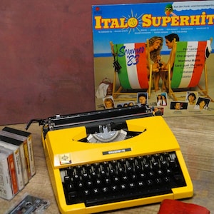 May include: A bright yellow typewriter with black keys and the brand name "BERETTE" is the focal point. Beside it are audio cassettes and a vintage "ITALO SUPERHITS" album cover. The album cover features a beach scene with the text "Sommer '83".
