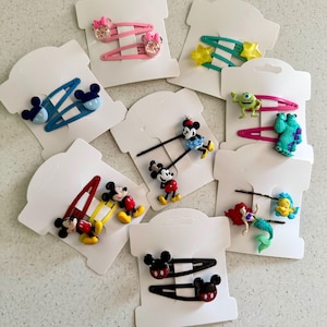 May include: A collection of colorful hair clips featuring cartoon characters. The clips include designs of Mickey and Minnie Mouse, stars, a green monster, and a mermaid. Each clip is attached to a white card.