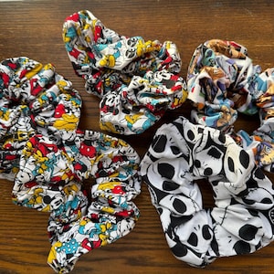 May include: Four colorful scrunchies with different patterns. One scrunchie has a white background with black and white skulls, another has a white background with colorful cartoon characters, and the other two have a white background with colorful cartoon characters.