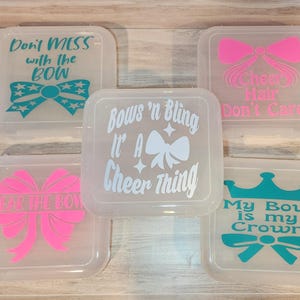 May include: Five clear plastic containers with lids, each featuring a different design related to cheerleading. The containers have text and bow graphics in various colors, including pink and teal. The text includes phrases like "Don't Mess with the Bow" and "Cheer Hair Don't Care."