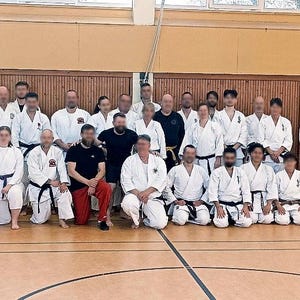 Könnte beinhalten: Gruppe von Personen in einer Sporthalle, die für ein Foto posieren. Die meisten tragen weiße Karate-Uniformen mit schwarzen Gürteln. Einige Personen sind in schwarzer Kleidung. Der Hintergrund umfasst eine Holzwand und einen polierten Holzboden.