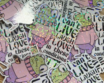 May include: Holographic stickers with a cartoon frog in a pink sweater and scarf. The stickers display the text "I can't LIVE LAUGH LOVE in these conditions."