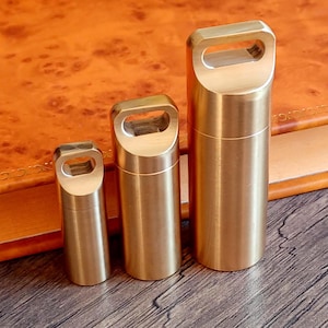 Op de afbeelding: Drie cilindrische messing containers van verschillende maten, elk met een rechthoekige handgreep. Ze zijn gerangschikt op een houten oppervlak, met een leren boek op de achtergrond. Het messing heeft een warme, gouden tint.