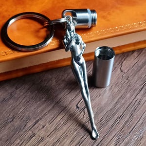 Puede incluir: Un llavero plateado con una figura femenina estilizada. El llavero incluye un anillo, un recipiente cilíndrico y una tapa roscada. Los artículos se muestran sobre una superficie de madera y un libro encuadernado en cuero.