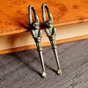 Può includere: Due cucchiai da portata antichi color bronzo con figure di scimmia. Sono appoggiati su una superficie di legno accanto a un libro rilegato in pelle. I cucchiai hanno un design dettagliato e un'estetica vintage.