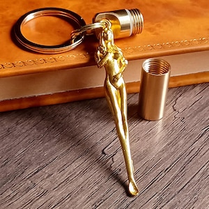 Può includere: Un portachiavi color oro con una figura femminile snella. Il portachiavi include un anello, un componente cilindrico e un tubo filettato separato. Gli oggetti sono esposti su una superficie di legno e un libro rilegato in pelle.