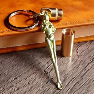 May include: A gold-toned metal keychain featuring a slender, standing figure charm attached to a circular ring. The charm includes a detachable cylindrical capsule with a threaded screw top, revealing a hollow interior for storage purposes.