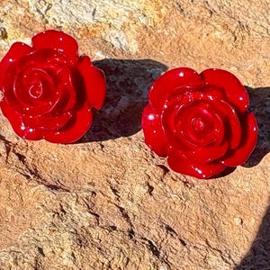 May include: Two glossy, bright red rose-shaped earrings. The earrings are set against a textured, tan-colored stone surface. The floral design is detailed, with visible petals and a central spiral.