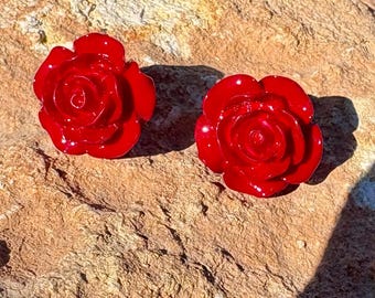 May include: Two glossy, bright red rose-shaped earrings. The earrings are set against a textured, tan-colored stone surface. The floral design is detailed, with visible petals and a central spiral.