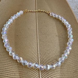 May include: A clear, iridescent beaded necklace with a gold-colored chain clasp. The necklace is composed of faceted beads that catch the light, creating a shimmering effect. The necklace is displayed on a gold-colored, textured surface.