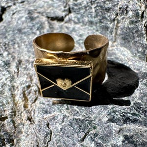 May include: A gold-toned open band ring featuring a rectangular black enamel plaque shaped like an envelope. The center of the envelope design displays a small, raised gold-colored heart detail. The ring band has a textured, uneven surface.