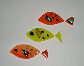 May include: Three glass fish ornaments, two orange and one yellow, with iridescent accents and black details. The fish are arranged in a row on a white background.