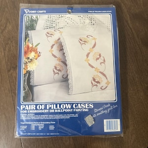 May include: A package of Vogart-Crafts pillow cases for embroidery or ballpoint painting. The pillow cases are white with a floral scroll design in brown and yellow. The package includes two pillow cases, each measuring approximately 20 inches x 32 inches.