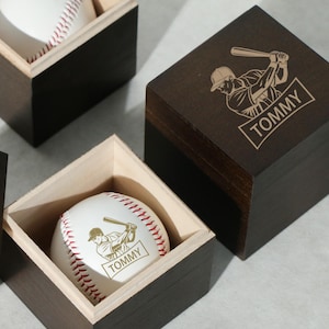 Op de afbeelding: Honkbal in een houten doos. De honkbal is wit met rode stiksels en een honkbalspeler-afbeelding met de naam "TOMMY". De doos is donkerbruin met een honkbalspeler-afbeelding op het deksel.