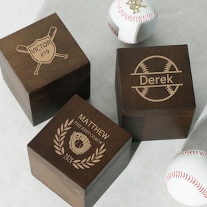 Könnte beinhalten: Drei dunkle Holzkisten mit Baseball-Gravuren. Eine Kiste trägt die Aufschrift "VICTOR #19", eine andere "Derek" und die dritte "MATTHEW THE BEST COACH 2026". Zwei Basebälle mit roten Nähten sind ebenfalls vorhanden.