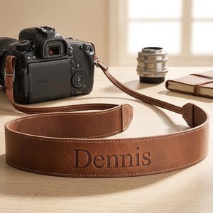 May include: A brown leather camera strap with white stitching and the name Dennis embossed in the center. The strap is attached to a black Canon DSLR camera, with a separate camera lens and a small brown notebook visible in the background.