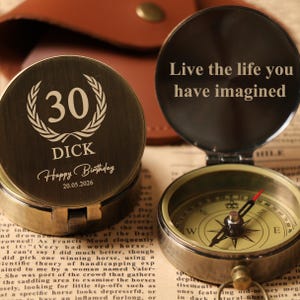 May include: A vintage-style compass with a bronze finish, open to reveal the directional dial. One side of the compass is engraved with "Live the life you have imagined." The other side is engraved with "30", "DICK", "Happy Birthday" and the date "20.05.2026".