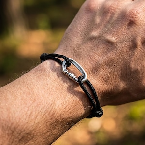 Peut inclure: Un bracelet en cordon noir doté d'un fermoir mousqueton en métal argenté. Le mousqueton présente une finition métallique lisse et porte l'inscription gravée 2026 sur le côté. Le cordon est tressé et fixé à travers les boucles du mousqueton.