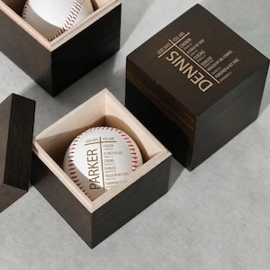 Peut inclure: Une balle de baseball dans une boîte en bois foncé. La balle est blanche avec des coutures rouges et le nom "PARKER" imprimé dessus. La boîte a un intérieur en bois clair et le texte "GOD SAYS YOU ARE" et d'autres textes imprimés sur la balle.