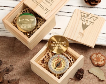 May include: Two square light-colored wooden boxes with sliding lids. One lid is engraved with 'Jasmine' and '16.07.2025'. The other lid features a mountain graphic and 'Marcus'. Inside, one gold compass is visible with the text 'I hope my gift can bring you good luck'.