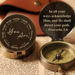 May include: Two open antique brass compasses with inspirational text. One compass reads "You Are" surrounded by positive affirmations. The other compass has a quote from Proverbs 3:6. The compasses are on a vintage paper background.