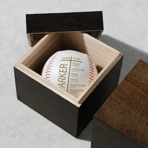 Puede incluir: Una pelota de béisbol con costuras rojas y el nombre "PARKER" impreso, dentro de una caja de madera. La pelota de béisbol tiene texto inspirador que incluye "CHOSEN" y "FEARLESS". La caja es de color marrón oscuro con un interior de madera más clara.