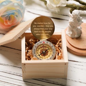 Peut inclure: Une boîte en bois ouverte contenant une boussole dorée avec des marquages noirs et rouges, posée sur du papier déchiqueté. Au-dessus, un disque doré porte l'inscription : « Peu importe où vous errez, puissiez-vous toujours retrouver le chemin vers ceux qui vous aiment ».