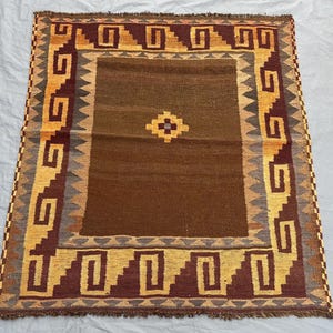 May include: A rectangular, hand-woven rug with a central brown field and a geometric border. The border features repeating patterns in shades of gold, brown, and gray. A diamond-shaped design is in the center of the rug.