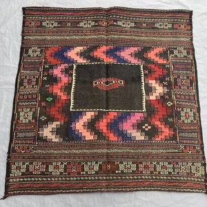 May include: A square, hand-woven rug with a central dark brown field and a geometric design. The rug features a border with repeating patterns in shades of red, brown, and cream. The central design includes red, pink, and blue zigzags.