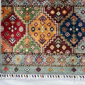 May include: A rectangular area rug featuring a geometric pattern of large diamonds in red, orange, green, and blue. The rug has a light blue border with small floral motifs and a fringe edge with multi-colored tassels along the bottom side.