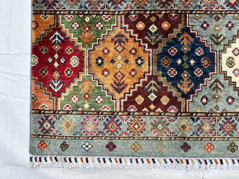 May include: A rectangular area rug featuring a geometric pattern of large diamonds in red, orange, green, and blue. The rug has a light blue border with small floral motifs and a fringe edge with multi-colored tassels along the bottom side.