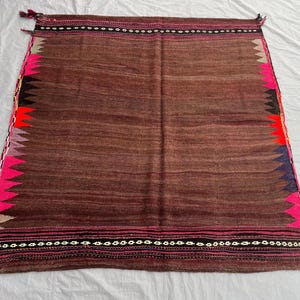 May include: A brown woven textile with a rectangular shape. The edges feature a zig-zag pattern in pink, black, red, and blue. The top and bottom edges have decorative stitching and tassels. The textile is likely a rug or decorative piece.