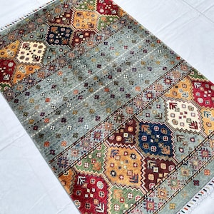 May include: A rectangular area rug featuring a central field of light gray with small repeating floral motifs, flanked by wide borders of geometric patterns in red, yellow, green, and blue, finished with short white fringe on both ends.