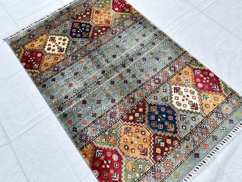 May include: A rectangular area rug featuring a central field of light gray with small repeating floral motifs, flanked by wide borders of geometric patterns in red, yellow, green, and blue, finished with short white fringe on both ends.