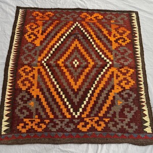 May include: Square, hand-woven rug with a geometric diamond pattern. The rug features a central diamond design in shades of orange, brown, and white, surrounded by a border of repeating patterns. The rug's edges are finished with a dark brown border.