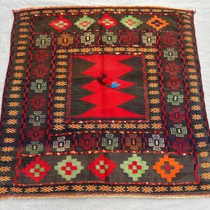 May include: A square, hand-woven rug with a central red geometric design. The rug features a dark brown background with repeating patterns in red, green, orange, and blue. The rug's border has a repeating geometric pattern.