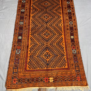 Pode incluir: Tapete retangular tecido à mão com um padrão geométrico em tons de laranja, castanho e azul marinho. O tapete apresenta um design central em diamante e uma borda detalhada com franjas. Dimensões: aproximadamente 91 x 152 cm.