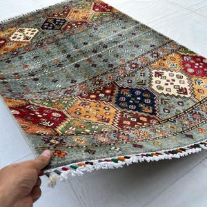 May include: A rectangular area rug featuring a sage green field with repeating geometric patterns in red, navy blue, yellow, and cream. The rug includes a series of horizontal bands with intricate floral and diamond motifs and white fringe.