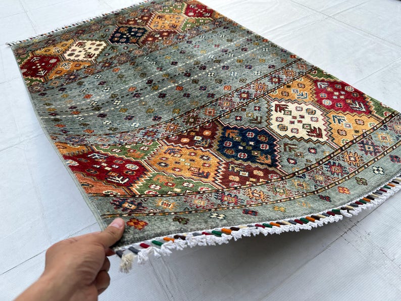May include: A rectangular area rug featuring a sage green field with repeating geometric patterns in red, navy blue, yellow, and cream. The rug includes a series of horizontal bands with intricate floral and diamond motifs and white fringe.