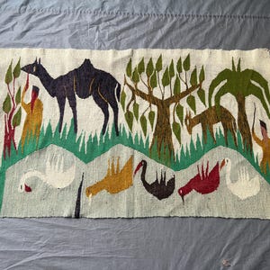 Puede incluir: Un tapiz rectangular tejido con fondo crema que muestra un camello oscuro, árboles verdes y un animal marrón. La sección inferior presenta una fila de cinco aves estilizadas en blanco, amarillo, negro y rojo sobre un campo de color verde claro.