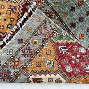 May include: A folded area rug featuring a complex geometric pattern with vibrant shades of mustard yellow, deep red, forest green, and slate gray. The rug displays intricate floral and diamond motifs, finished with white fringe along the edge.
