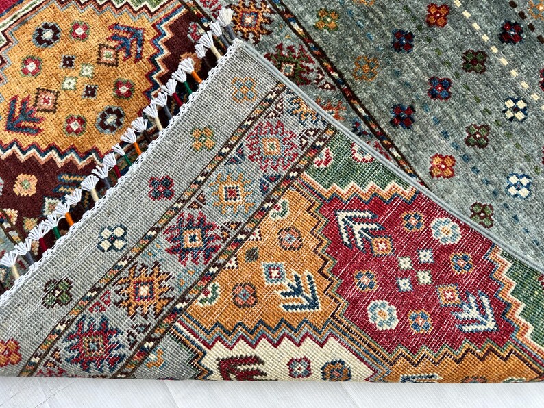 May include: A folded area rug featuring a complex geometric pattern with vibrant shades of mustard yellow, deep red, forest green, and slate gray. The rug displays intricate floral and diamond motifs, finished with white fringe along the edge.