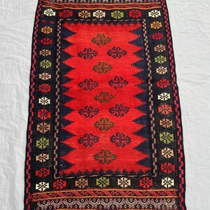 May include: A rectangular, hand-woven rug with a red central field and a dark blue border. The rug features geometric patterns in various colors, including black, green, and gold. The rug's dimensions are not visible.