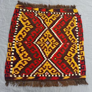 May include: A rectangular, hand-woven rug with a geometric pattern. The rug features a central diamond design in red, yellow, and brown, with white accents. The edges are finished with a brown fringe. The rug measures approximately 24 inches by 24 inches.