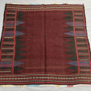 May include: A rectangular, hand-woven rug with a deep burgundy center and intricate borders. The borders feature red, black, blue, and white geometric patterns. The rug's design includes repeating triangular shapes and a fringed edge.