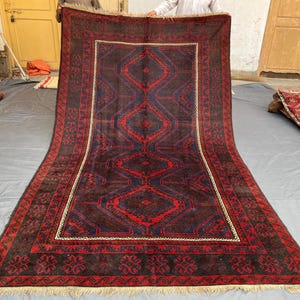 Pode incluir: Um tapete retangular grande com um design geométrico central em vermelho e azul sobre um fundo marrom escuro. O tapete tem uma borda com padrões florais repetidos e uma franja creme.