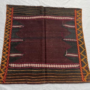 May include: A rectangular, hand-woven rug with a central maroon field. The rug features black and orange horizontal stripes, geometric patterns, and a zig-zag border in orange, red, and yellow. The rug has a distressed appearance.